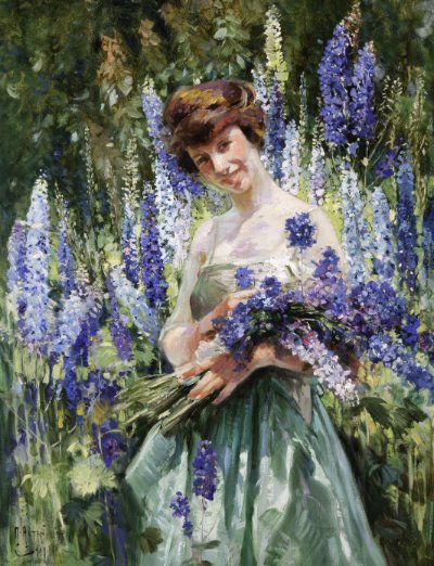 An image of a woman with brunette hair in an updo. She is wearing a pale green dress. She is holding freshly cut delphinium flowers, which are in shades of blue and purple. She is standing in front of a background of delphiniums and leaves.